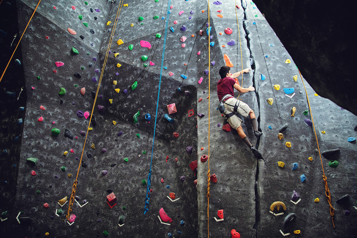 A student climbs an indoor rock wall using ropes and climbing holds.