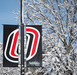 A campus banner with the University of Nebraska at Omaha logo hangs on a lamppost. Snow-covered trees fill the background under a bright blue sky. The banner features the UNO logo in red, white, and black.