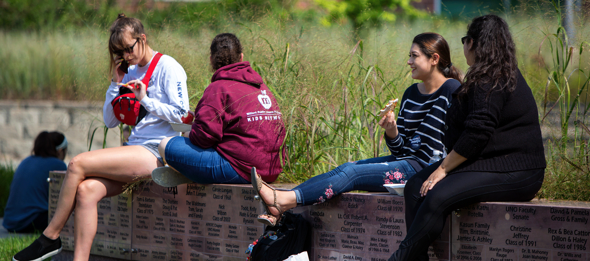 Five college students sit on a low brick wall outdoors, eating lunch and talking on a sunny day with backpacks at their feet.