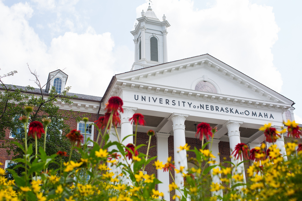 Low-angle view of the University of Nebraska at Omaha building with white columns and a cupola, framed by red and yellow flowers in the foreground under a partly cloudy sky.