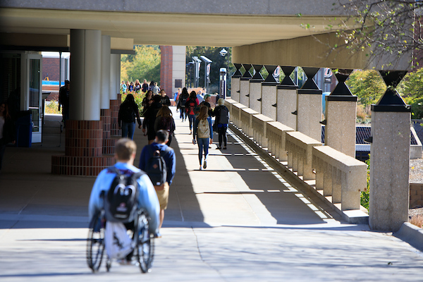 A student using a wheelchair travels along a covered campus walkway with other students walking nearby.