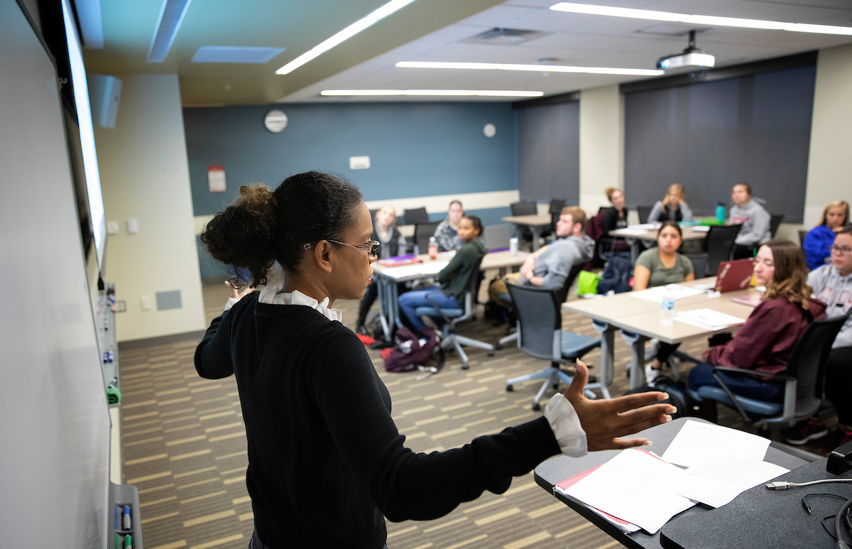 An instructor gestures while teaching at the front of a classroom as students sit at tables and listen during a lecture.