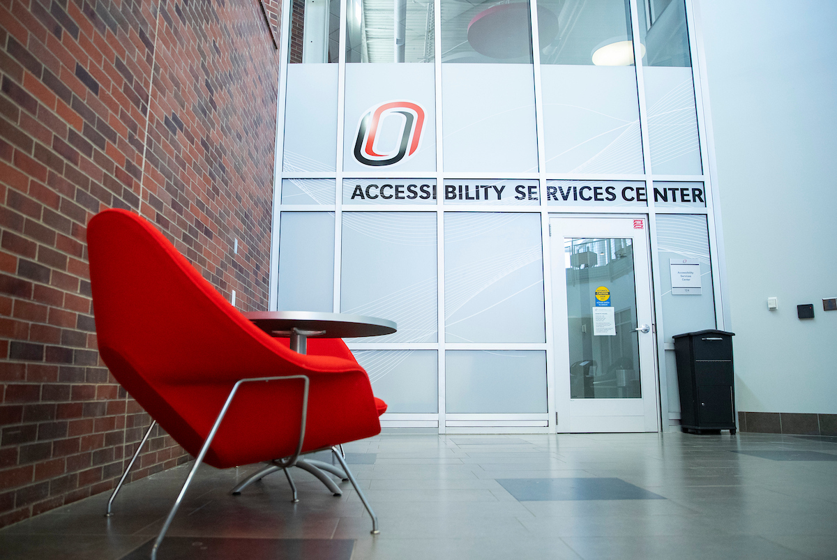 Interior of a university building with a glass-walled entrance labeled “Accessibility Services Center,” featuring a large red chair and small round table in the foreground, brick wall on the left, and a glass door with posted notices on the right.
