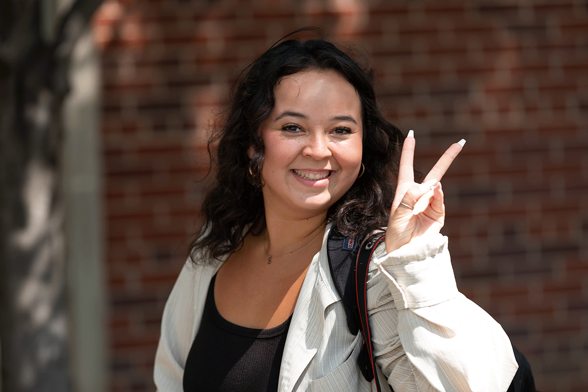 A smiling student with a backpack stands outdoors in front of a brick wall, holding up a peace sign with her hand.