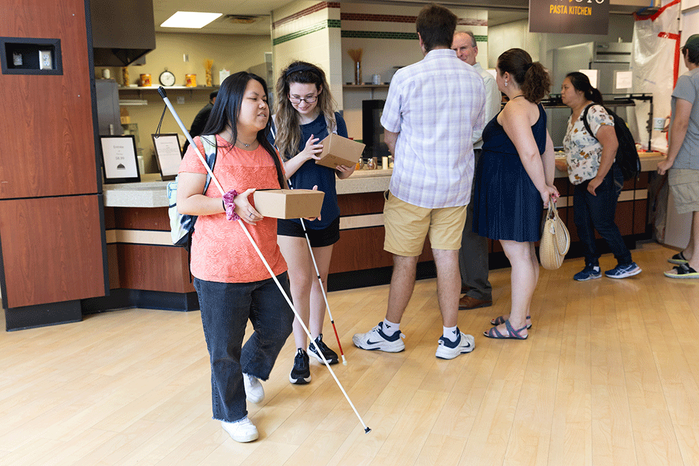 A student using a white cane walks through a campus dining area carrying a takeout container, while other students stand in line at a food counter in the background.