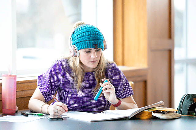 a student wearing a teal beanie sits at a table studying.