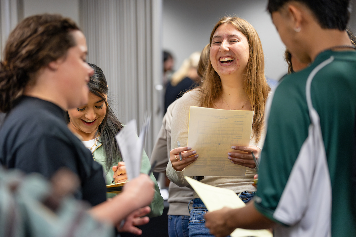 A group of students stand together indoors, smiling and reviewing papers while chatting in a casual, collaborative setting.