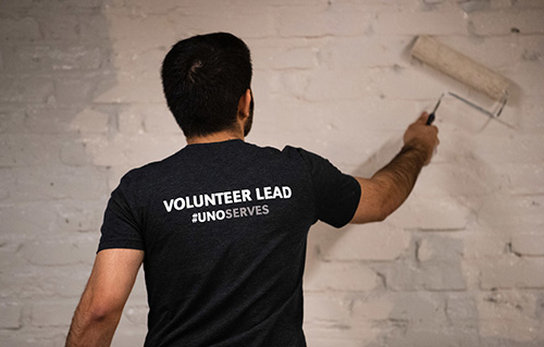 A student wearing a "Volunteer Lead, UNO Serves" t-shirt rolls paint onto the wall.