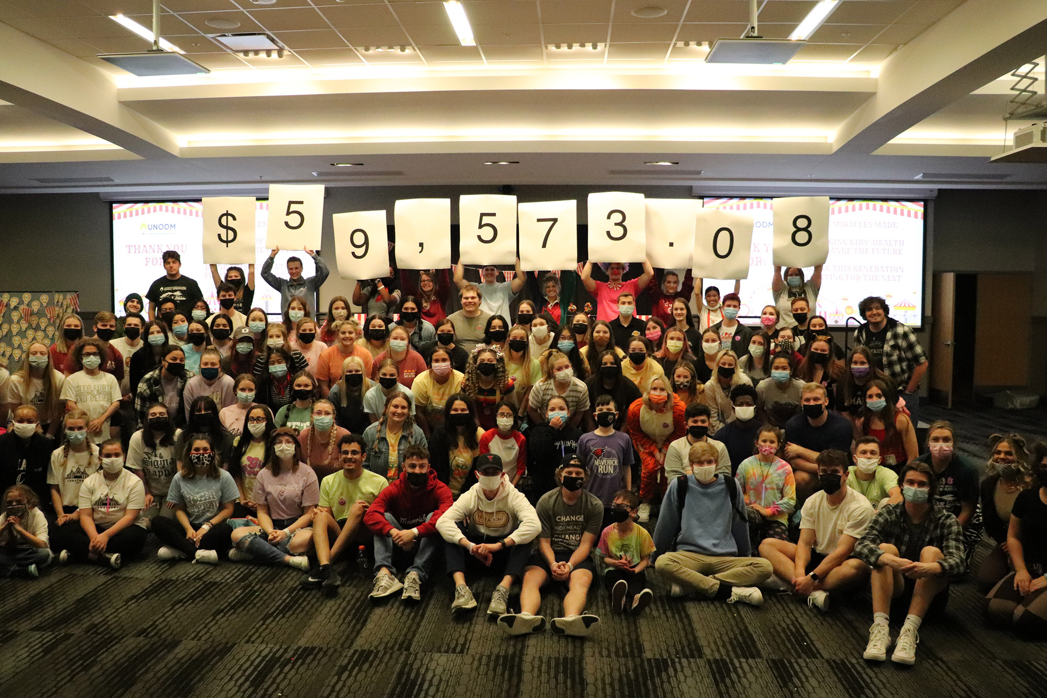 Large group of masked students posing indoors while people in the back hold placards spelling out “$59,573.08.”