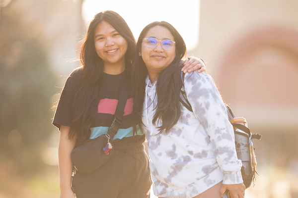 Two students stand close together, smiling warmly at the camera in soft sunlight. One student wears a black shirt with colorful stripes, and the other wears a tie-dye hoodie and carries a backpack. The background is softly blurred, creating a bright and friendly atmosphere.